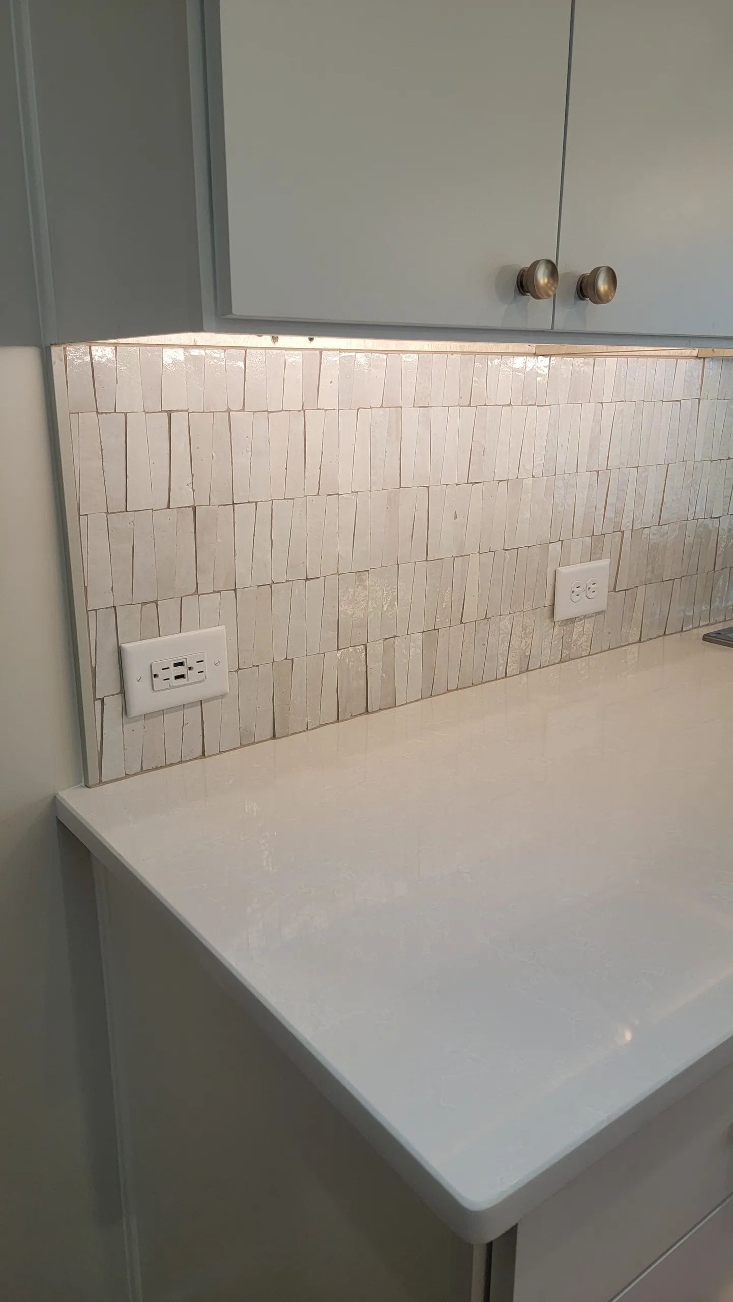 White glossy stacked-tile kitchen backsplash above a quartz counter and under-cabinet lighting, with outlets and cabinets