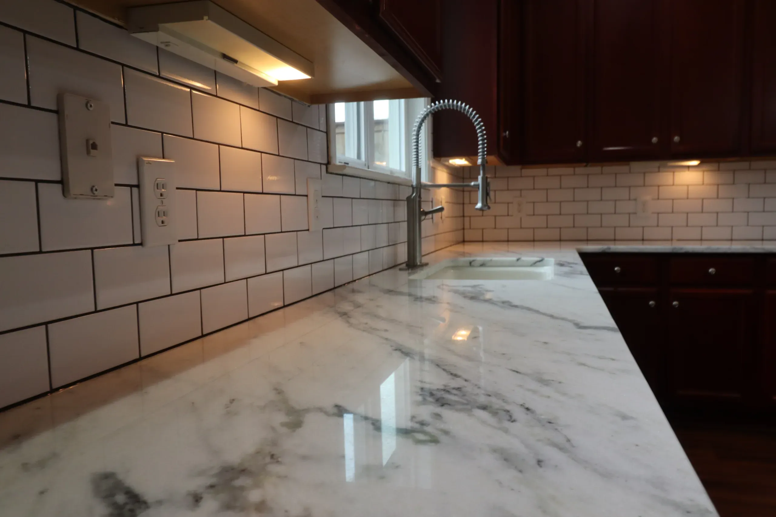 White subway tile kitchen backsplash with dark grout, marble countertop, and farmhouse sink under a window