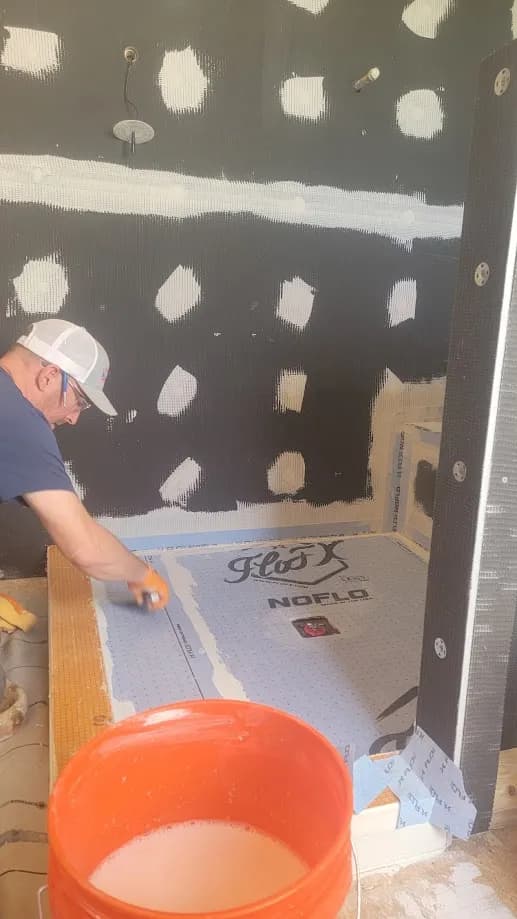Worker installing shower pan liner during tile repair, with patched walls and orange bucket in foreground